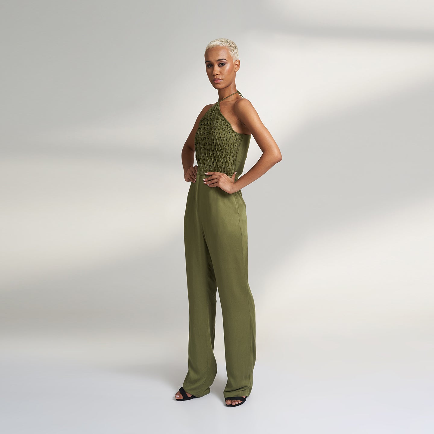 A global small size model wearing a solid olive green jumpsuit, highlighted with hand-stitched smocking on the bodice panel with a drawstring attached to the bodice to tie at the neck. inspired from the vintage window designs.