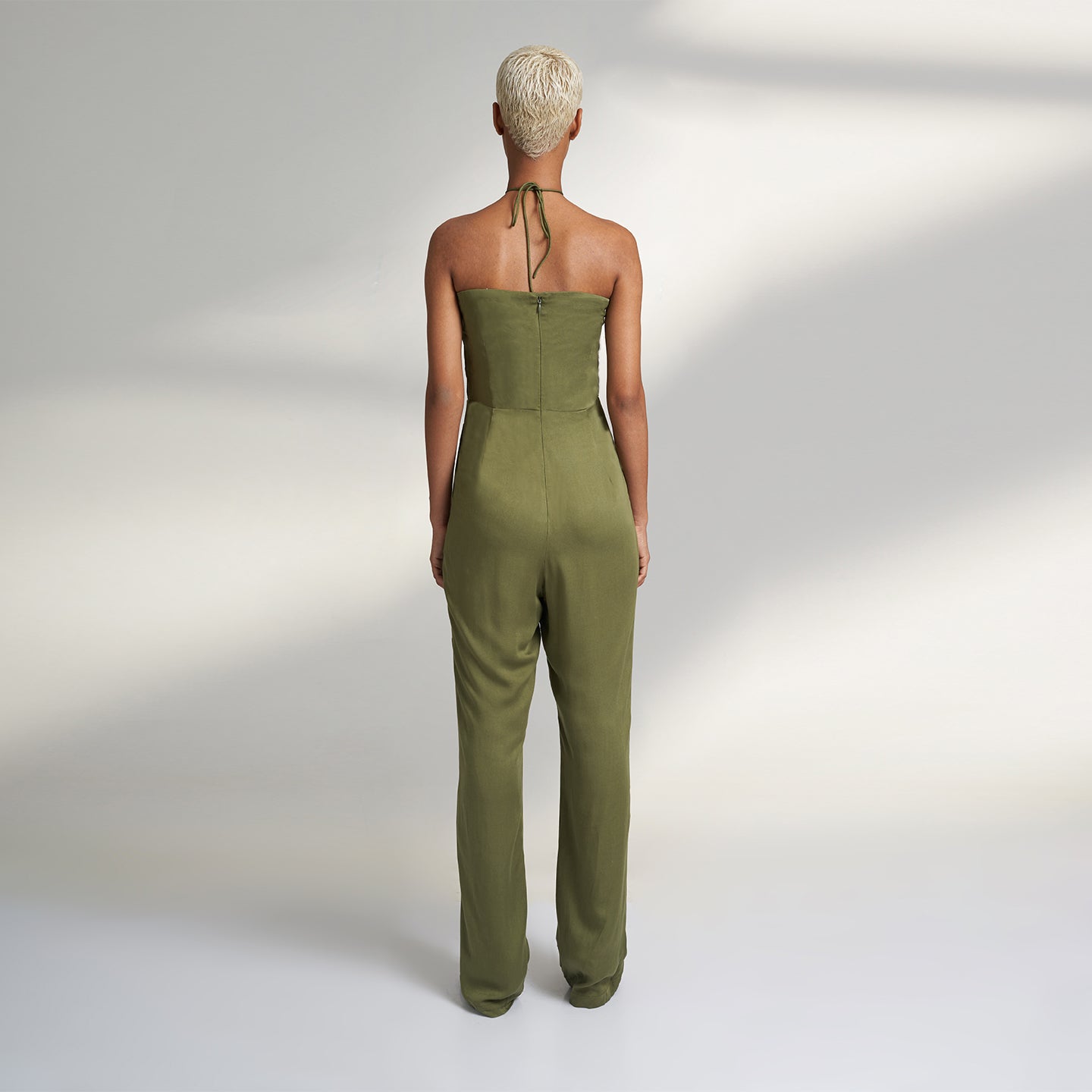 A global small size model wearing a solid olive green jumpsuit, highlighted with hand-stitched smocking on the bodice panel with a drawstring attached to the bodice to tie at the neck. inspired from the vintage window designs.