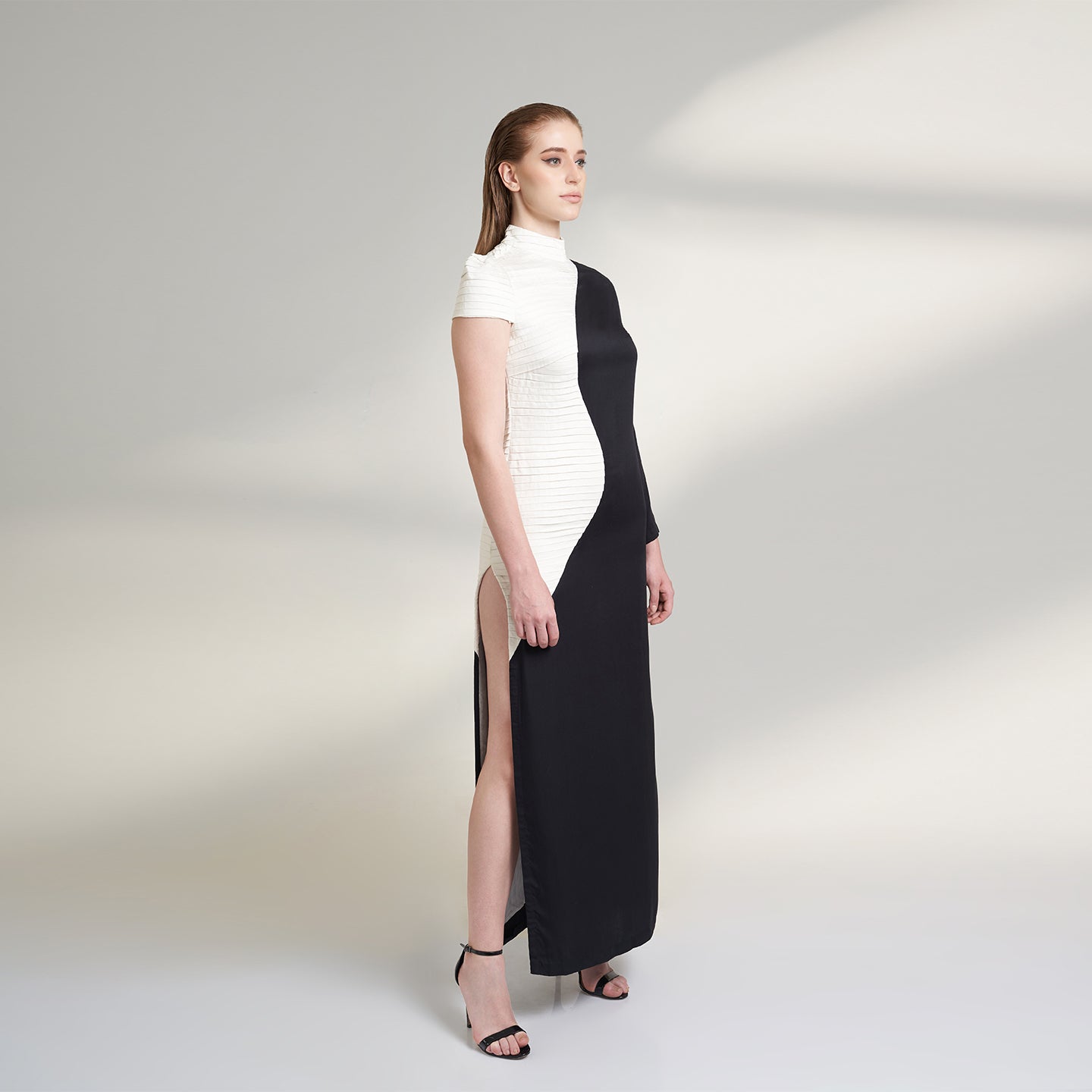 A medium size model wearing a black and white half and half dress made from organic lotus stem fabric. This sustainable vegan dress is a long floor length dress with one side pleated in white and one side plain black with a thigh high side slit striking a side pose.