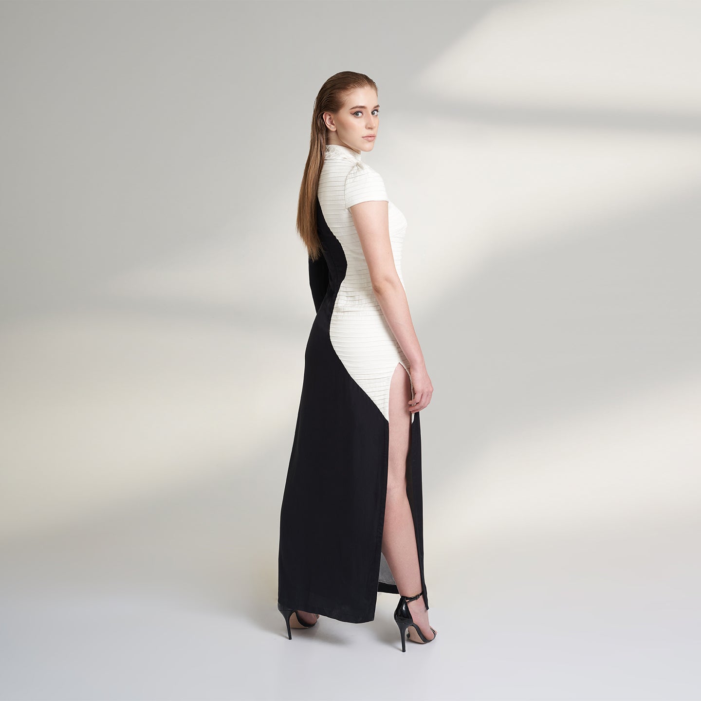 A medium size model wearing a black and white half and half dress made from organic lotus stem fabric. This sustainable vegan dress is a long floor length dress with one side pleated in white and one side plain black with a thigh high side slit striking a side pose.
