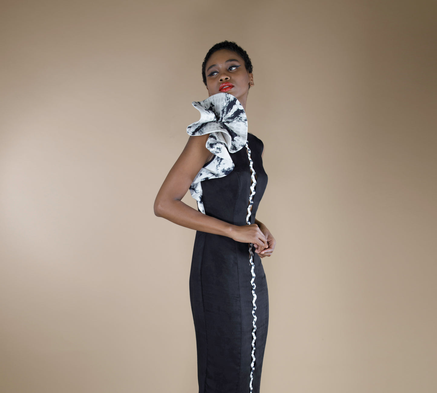 A global small size model wearing an organic black dress with tonal print in calf length black dress with a zig zag centre front panel with aztec print in black and white and a 3D flower motif on shoulder made from organic lotus stem fabric, striking a side pose.